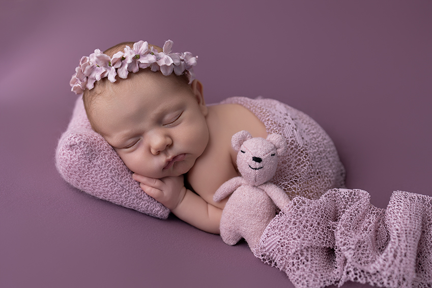 Sleeping newborn baby photographed in a warm studio in Royston, Hertfordshire on purple with floral headband and teddy
